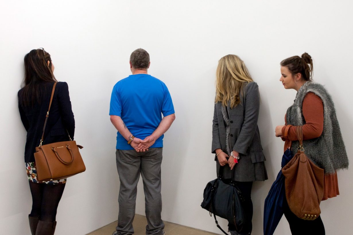 A middlA middle-aged man in casual clothing stands, hands clasped behind his back, facing the corner of a bare, white room. Three young people stand, watching the man.e-aged man in casual clothing stands, facing the corner of a bare, white room. Three young people stand, watching the man.