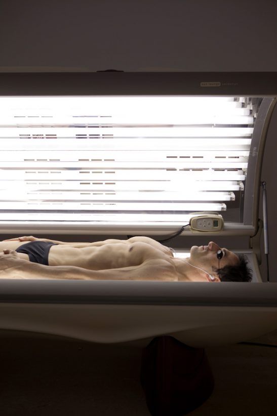 A young man with an athletic, muscular build, wearing black Speedos, black swimming goggles and small white earphones, lies in a glowing, electric sun bed.