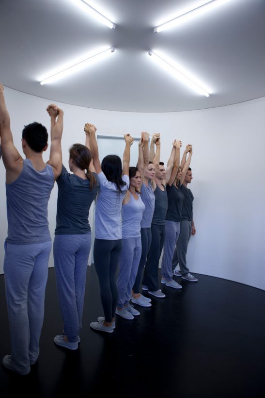 Eight young people stand in a line – five facing forwards, and three facing the opposite direction, in a small, bare, white circular room. The people are dressed in similar grey T-shirts, tank tops and sweat pants with matching grey shoes, arms upstretched, holding hands, with serious, focused expressions.