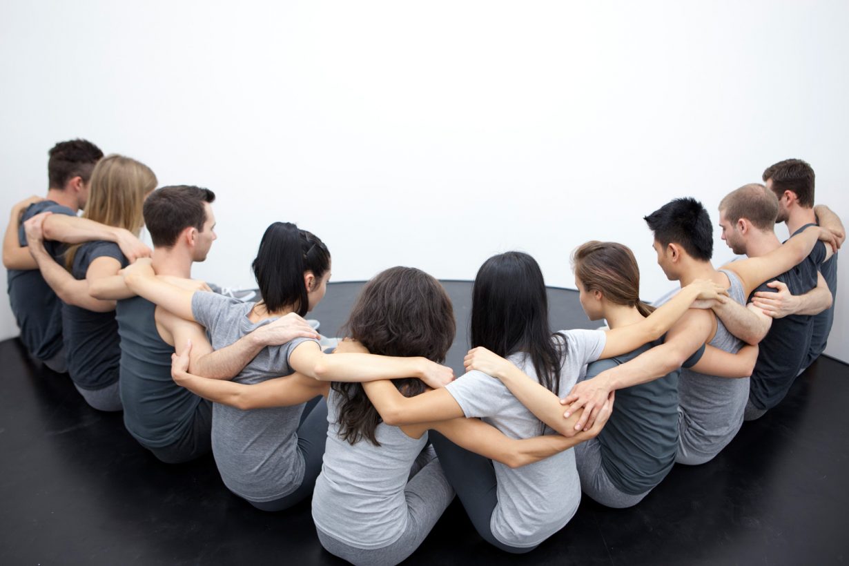 Ten young people sit in a V-shaped line, dressed in similar grey sportswear, with arms clasped around each other’s shoulders, in a bare, white room with a black rubber floor.
