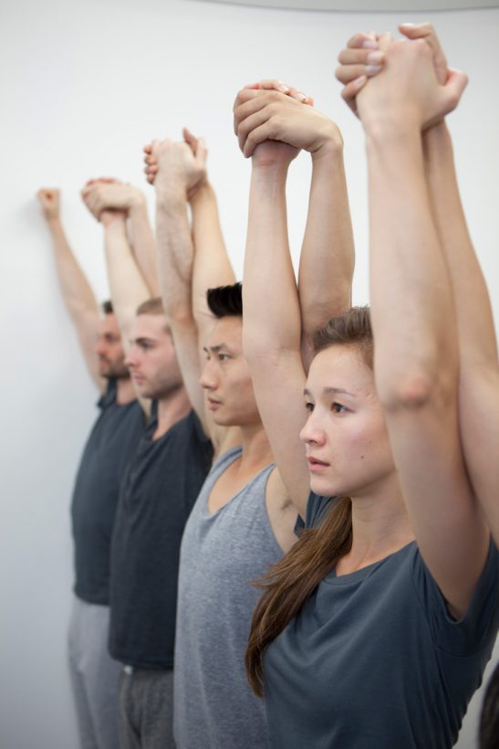 Five young people stand in a line, dressed in similar grey sportswear, arms upstretched, holding hands, with serious, focused expressions, in a bare, white room.