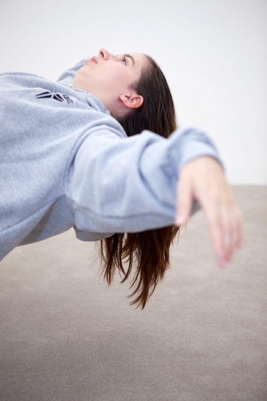 A young woman in loose, casual clothing leans back horizontally, in an impossibly balanced position, arms outstretched, in a bare white room.