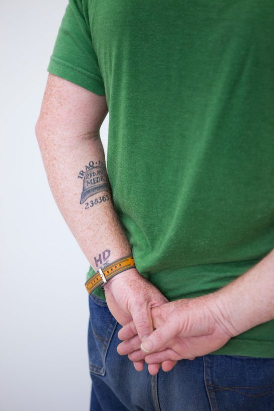 A middle-aged man in a green T-shirt stands, with his hands clasped behind his back, facing the corner of a bare, white room. On his arm is a tattoo featuring the words “Iraq” and “medic” and the image of a helmet.