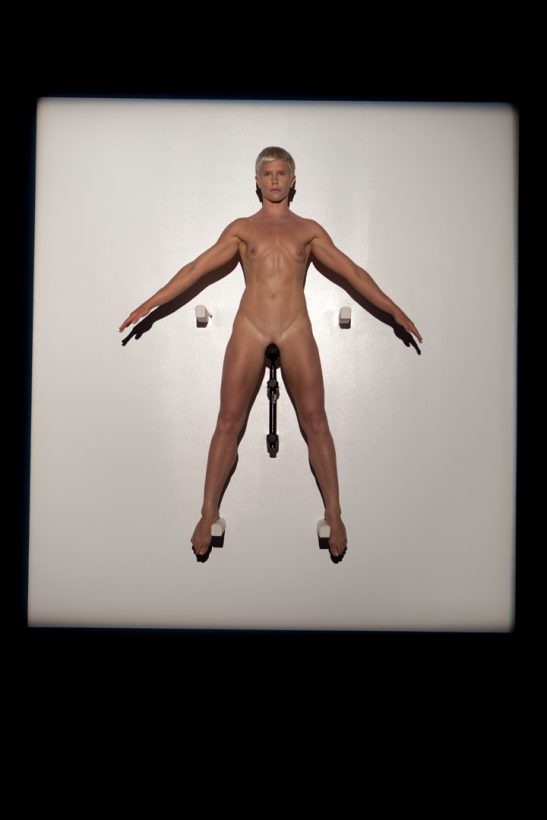 A naked woman sits, balancing, arms outstretched, on a bicycle seat, mounted high on a wall, in a glowing square of white light.