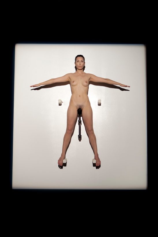 A naked woman sits, balancing, arms outstretched, on a bicycle seat, mounted high on a wall, in a glowing square of white light.