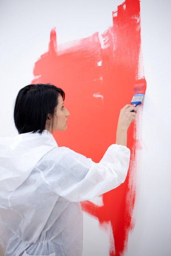A young woman in protective overalls paints a bare white wall with red paint.