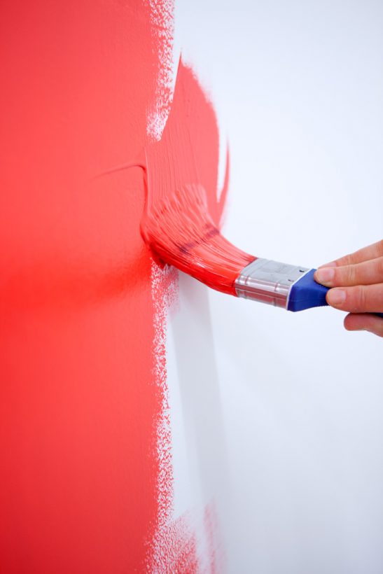 A hand holding a paintbrush paints a bare white wall with red paint.