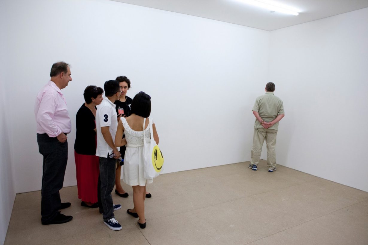 A middle-aged man in casual clothing stands, facing the corner of a bare, white room. A diverse group of five people stand in the opposite corner, in conversation, looking at the man.