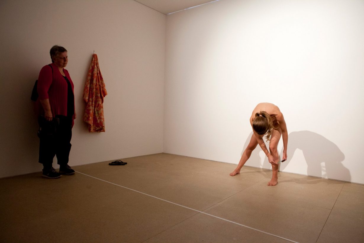 A naked young woman leans over and holds a small hand mirror against her leg. A robe hangs on a hook on the wall, with a pair of flip flops beneath. An older woman watches the young woman, in a bare white room.