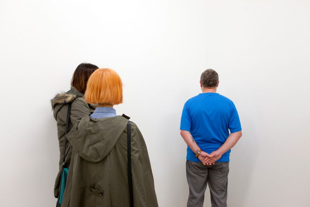 A middle-aged man in casual clothing stands, hands clasped behind his back, facing the corner of a bare, white room. Two young people stand, watching the man.