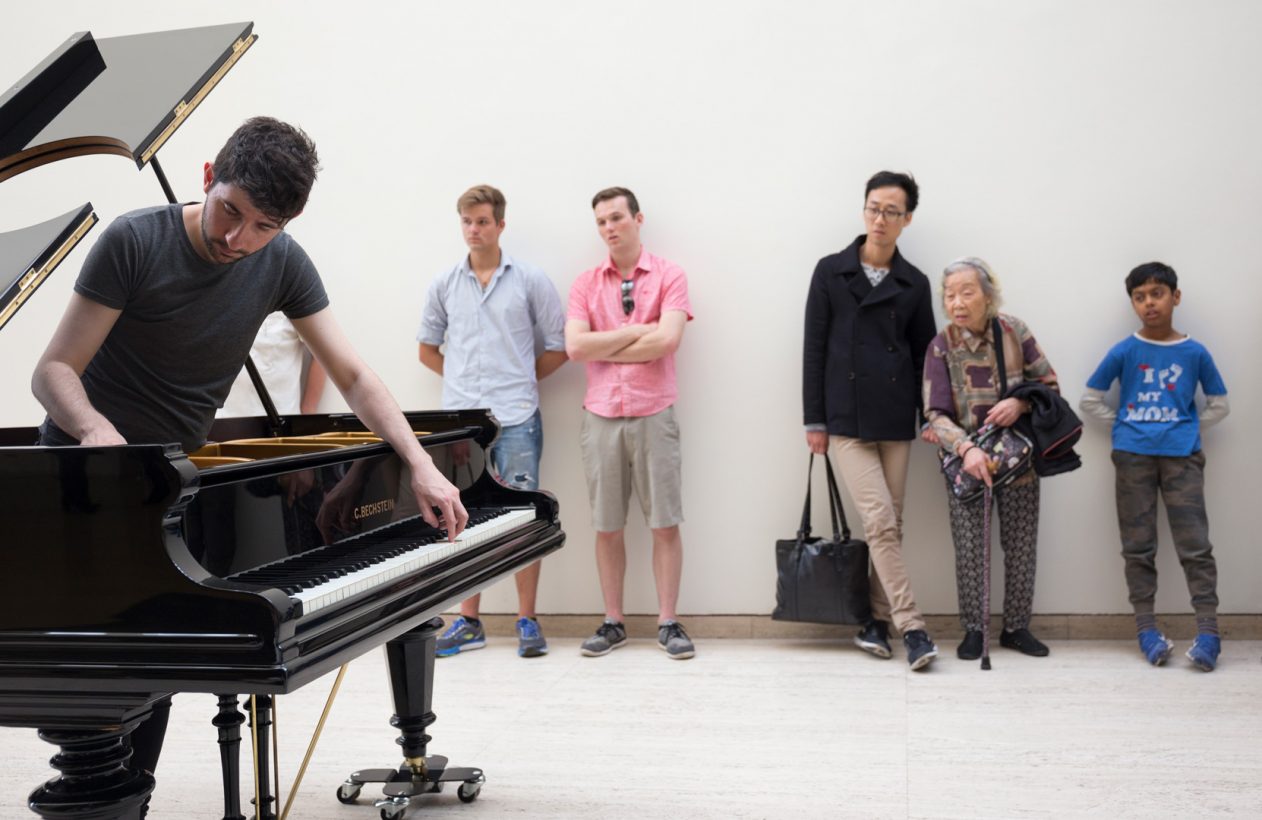 A man stands inside a grand piano, which has a round hole cut in its lid. The man plays the piano keys with one hand and plucks the strings with the other hand, with a look of intense concentration. Around 7 people stand against a bare white wall, watching the pianist.