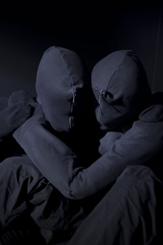 Two human figures, dressed in black, with their faces and hands entirely covered in black fabric, sit on the floor, leaning against each other, with arms intertwined, in a bare, darkened room.