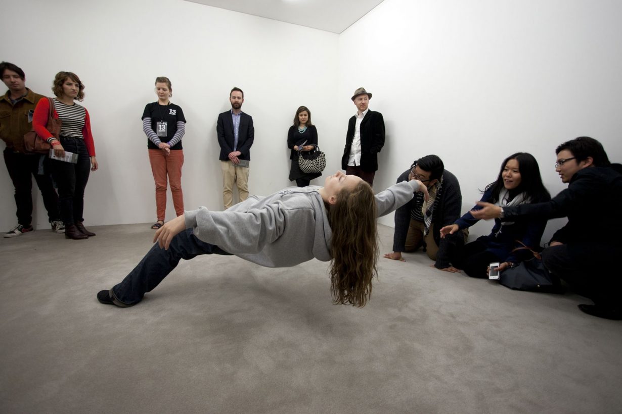 A young woman in loose, casual clothing leans back in an impossibly balanced position, with her feet on the floor, torso horizontal and arms outstretched. 9 people stand or crouch, watching the woman, in a bare white room.