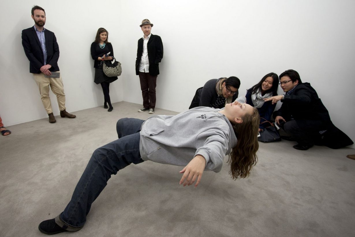 A young woman in loose, casual clothing leans back in an impossibly balanced position, with her feet on the floor, torso horizontal and arms outstretched. 6 people stand or crouch, watching the woman, in a bare white room.