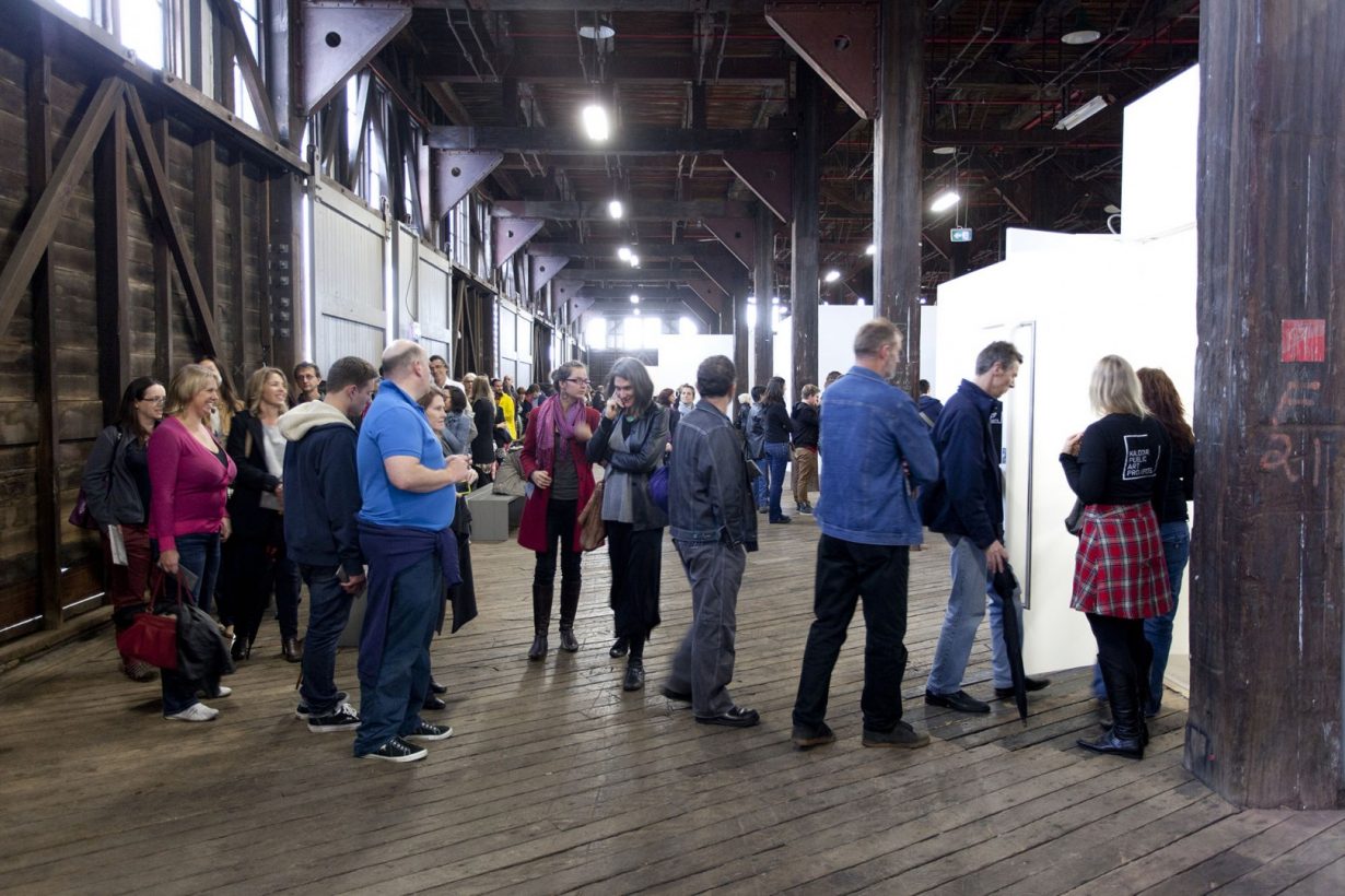 A crowd of around 25 people queues outside a small white room inside a large industrial space.