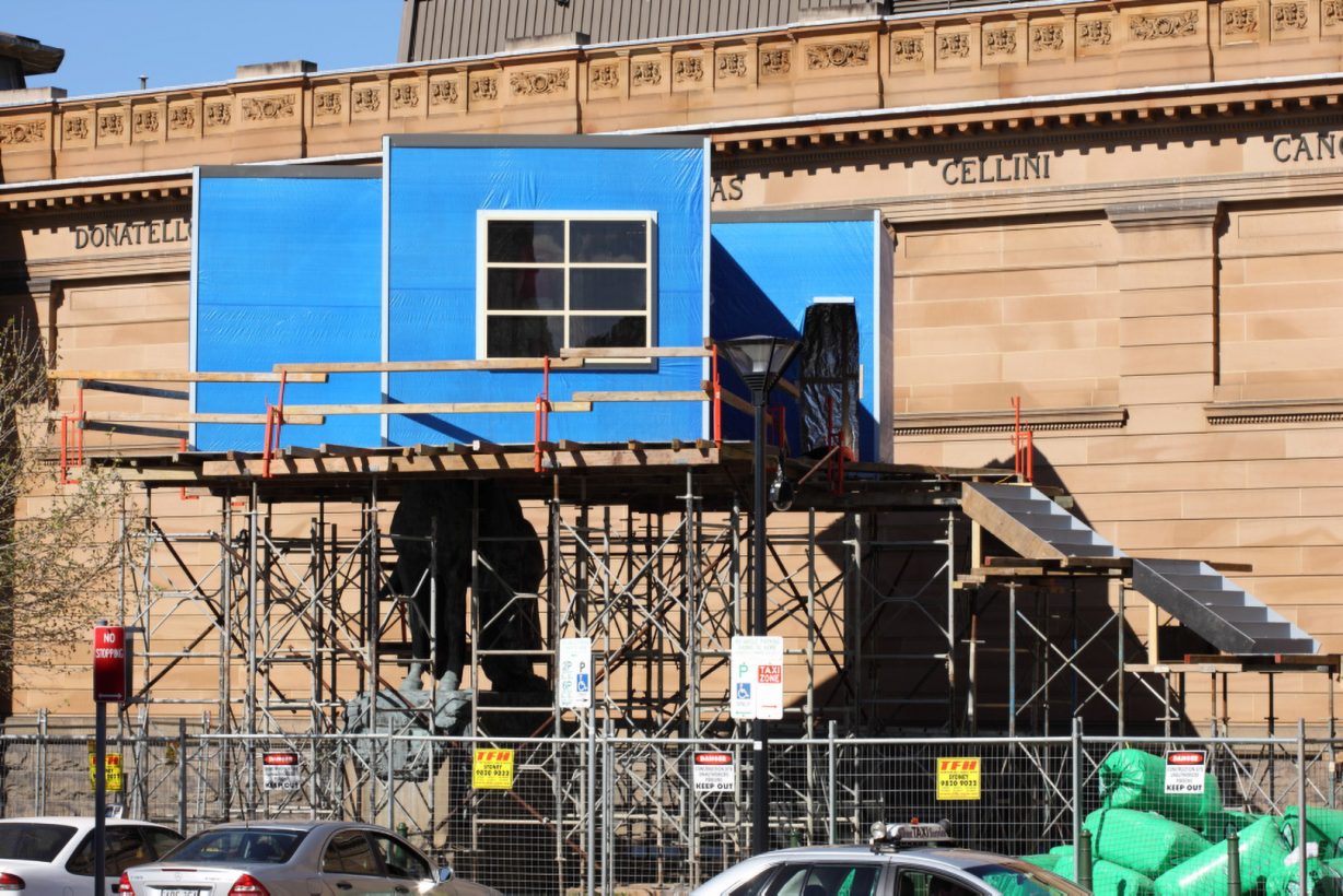 A blue box-like structure with a window, on top of a scaffolding base, with stairs, and surrounded by temporary fencing. The structure sits in front of a neo-classical sandstone building.