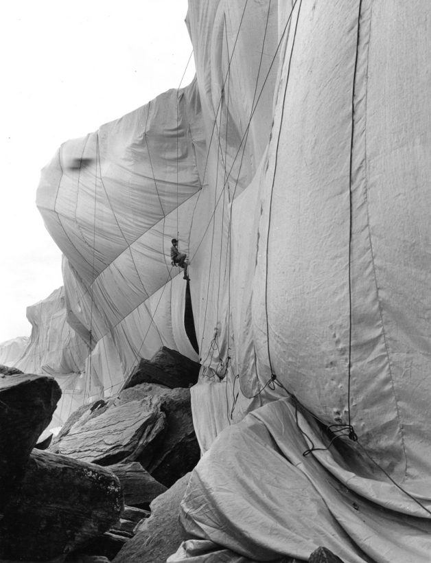 A man abseils down a large, craggy seaside cliff, which is wrapped in light-coloured fabric and rope.