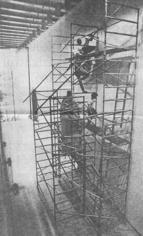 Two young people stand on three levels of scaffolding, against an art gallery wall.