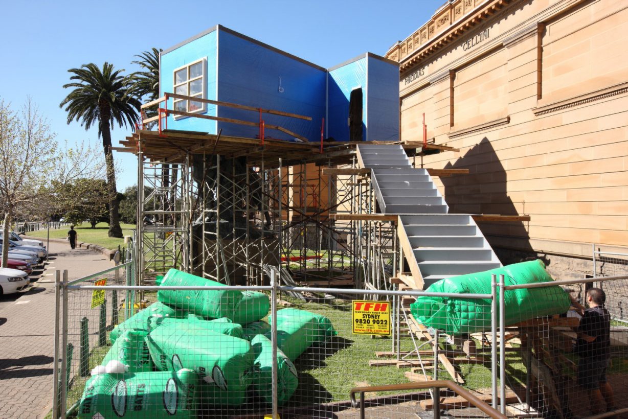 A blue box-like structure with a window, on top of a scaffolding base, with stairs, and surrounded by temporary fencing and construction materials. The structure sits in front of a neo-classical sandstone building.