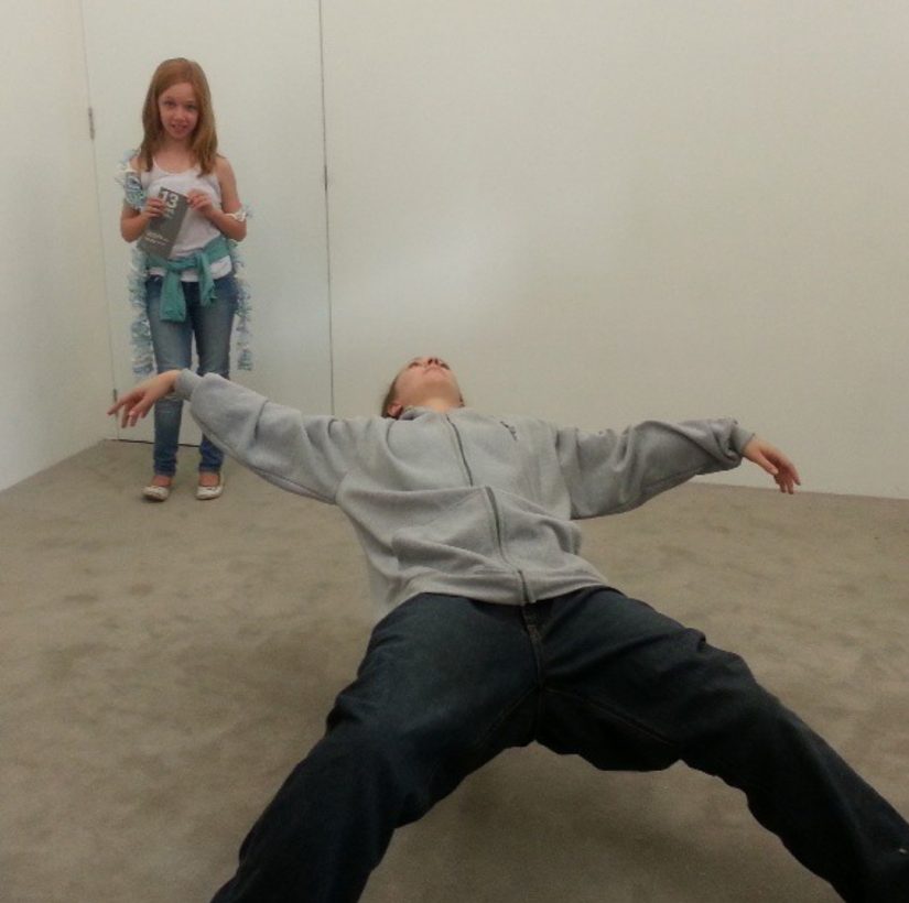 A young woman in loose, casual clothing leans back in an impossibly balanced position, with her feet on the floor, torso horizontal and arms outstretched. A 12-year-old girl stands, watching the woman, in a bare white room.