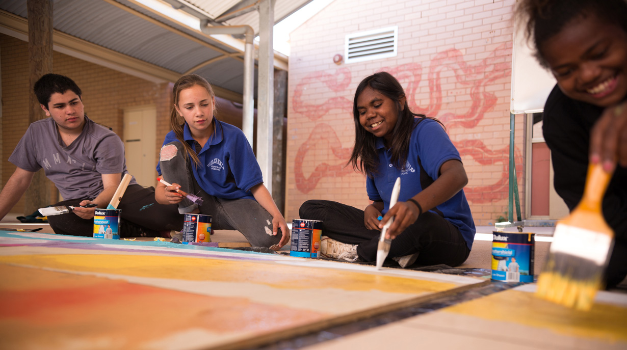 Four teenaged children sit on the floor, smiling, and painting with large paintbrushes and white and yellow paint onto a large timber board laid in front of them.