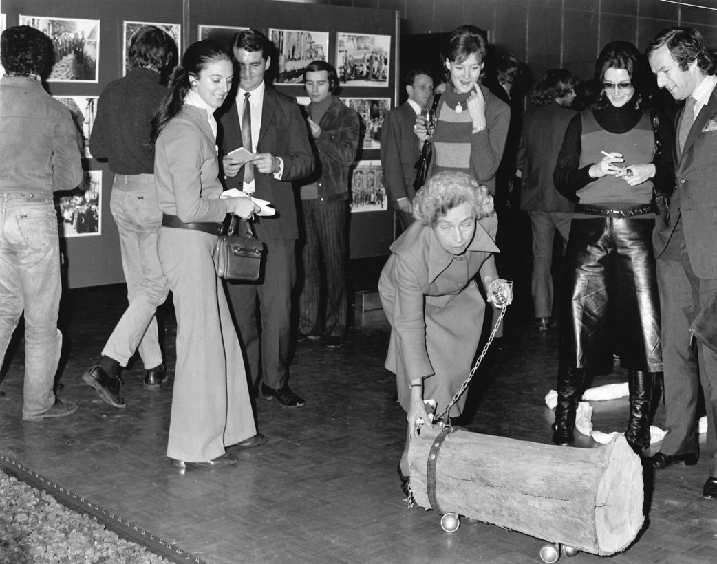 A group of people in fashionable 1970s clothing stand in an art gallery. The people are smiling, looking down at a small sawn-off log, with wheels and a dog collar. One older woman holds a chain attached to the collar, and crouches down to touch the log.