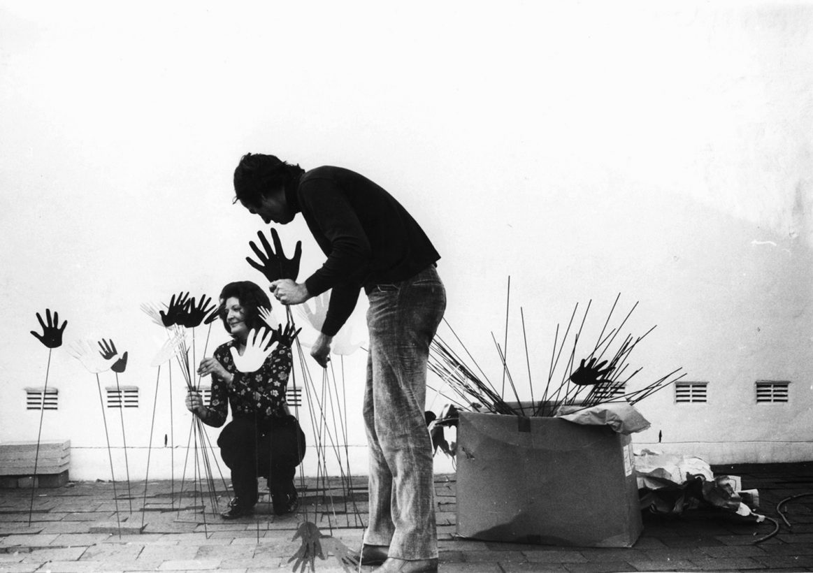A young woman, wearing a 1970s patterned shirt, and a man arrange a series of hand-shaped objects on wire stands.