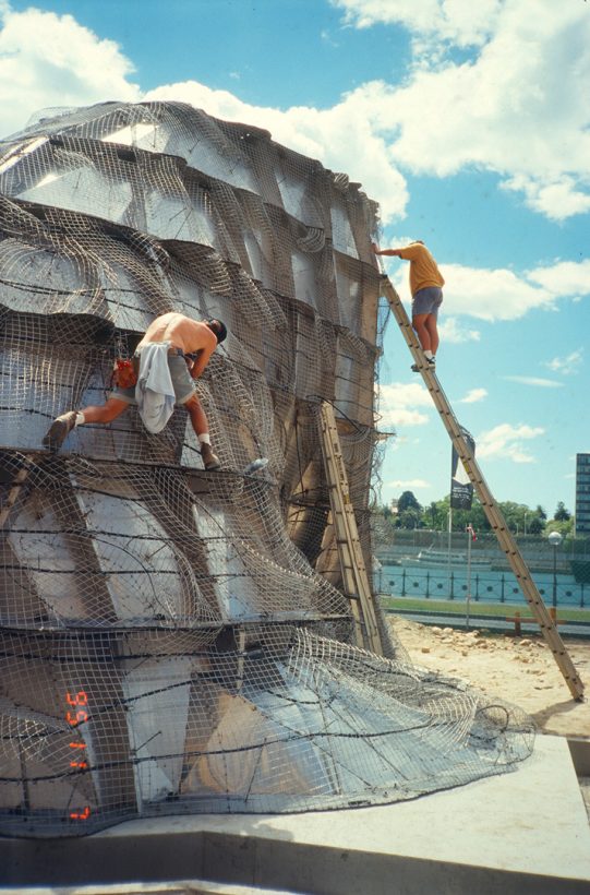 A young man, wearing work boots and no shirt, works while climbing on a large steel, mesh-covered structure. A second man stands on a ladder, leaning against the structure.