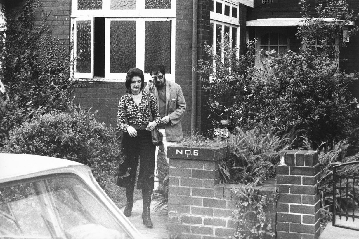 A young woman, in a fashionable 1970s outfit with patterned shirt, walks with a bearded man through the gate of a house towards a parked car.