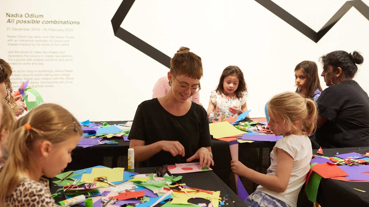A young woman sits at a table, smiling, and making a collage of coloured paper. A group of around 6 children and 3 adults sits around 3 tables, cutting coloured paper. The tables are covered in black cloth, and littered with coloured paper, scissors and glue. Text on the wall of the room reads, “Nadia Odlum, All possible combinations.”