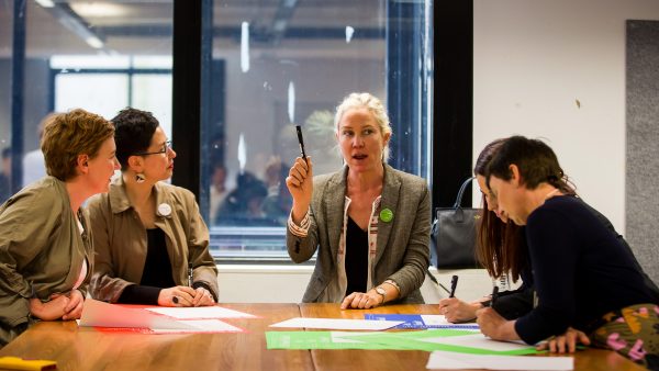 Five women sit at a large table, in conversation. One woman holds up a black marker and speaks, while two women listen intently. In front of the women are scattered red, green and blue posters. Two of the women make notes with black marker on posters.