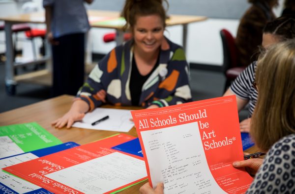 Three women sit at a large table, in conversation. One woman speaks, smiling broadly. One woman holds a red poster with the heading “All Schools Should be Art Schools”. The third woman writes notes with black marker on a blue poster.