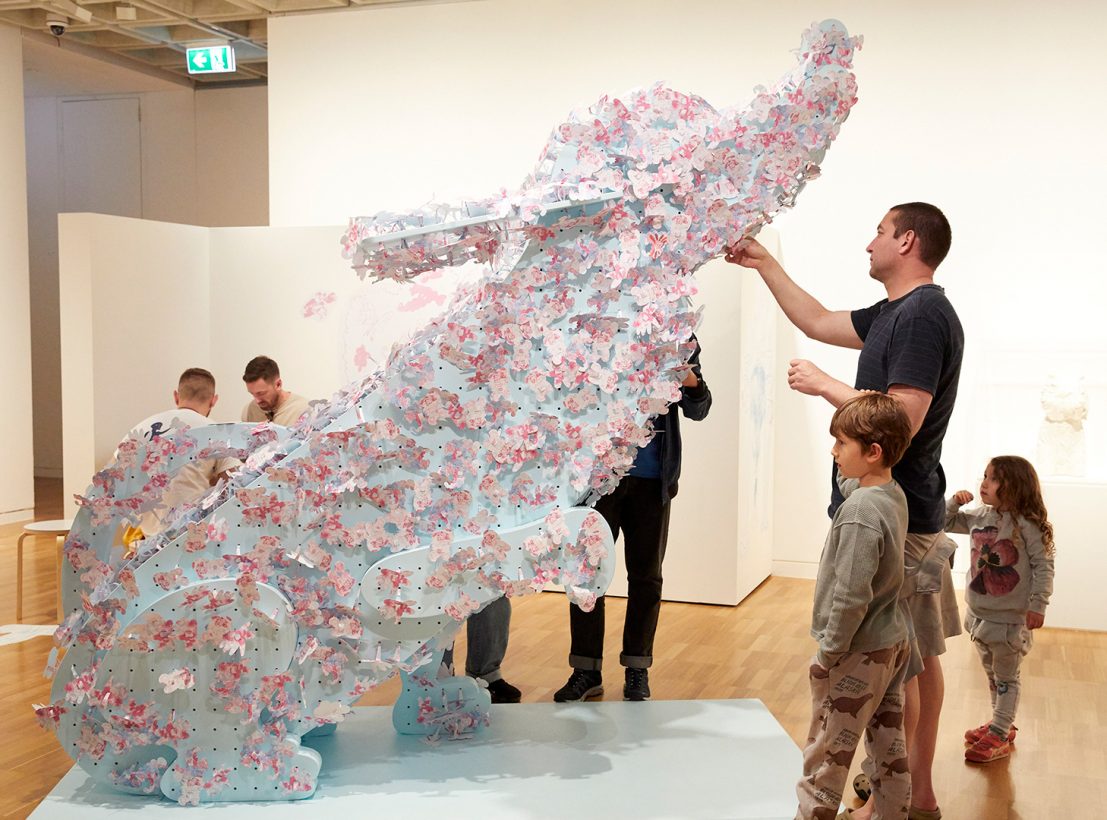A large, pale blue timber sculpture of a cartoon-like dog, sits, with one paw raised, in a modern art gallery. The dog is decorated with blue and pink paper flowers. A man and two small children stand nearby, as the man attaches a paper flower to the sculpture.