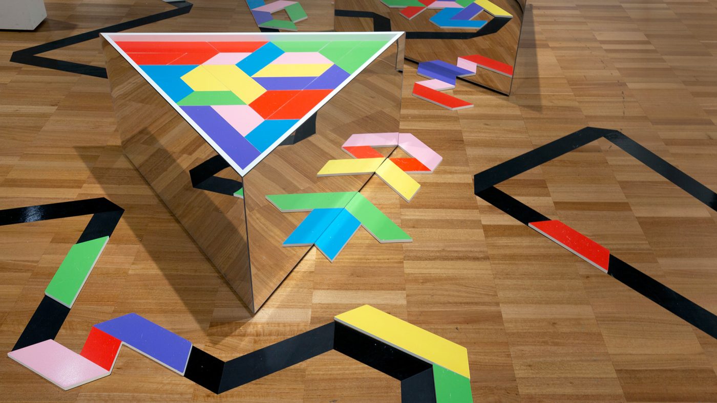 Two triangular mirrored tables, topped with an arrangement of geometric coloured blocks. A black line zig-zags across the floor.