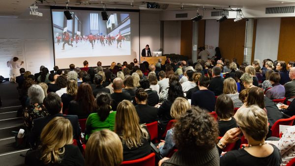 A man stands at a podium, speaking into a microphone, in an auditorium with a seated audience of around 200 people. A projected slide shows an image of school children running, with placards, in an art museum.