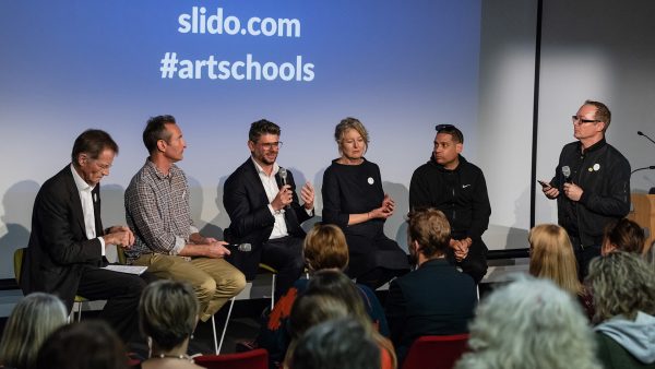 Four men and one woman, all dressed in business attire, sit in front of a seated audience. One of the men speaks into a microphone. A man stands beside the group, holding a microphone. Projected on a screen behind the group is the text “# art schools”